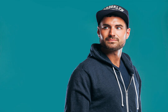 Attractive young man in stylish cap and hoodie looking away while standing over blue background at studio.