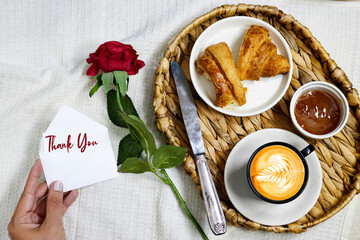 morning concept - Fresh baked croissant for breakfast, apricot jam and coffee cup with leaf late art on bed with rose flower and woman hand holding an enveloppe with thank you text 