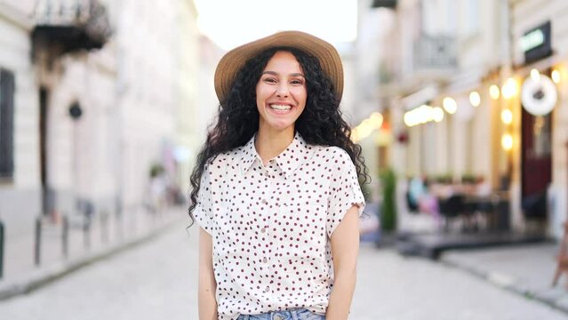 Portrait beautiful happy young tourist woman in a hat standing on a city street on urban town background. Joyful pretty traveler curly brunette smiling and looking at camera. Outdoor outside summer