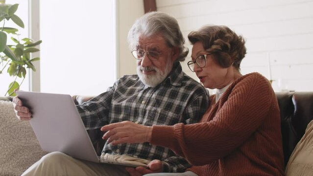 Middle aged older retired family couple using computer applications, having fun web surfing internet, shopping online or communicating distantly relaxing together on cozy sofa in living room.