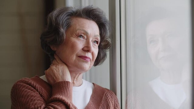 Thoughtful retired mature woman in casual clothes standing by large window. People, social distancing and quarantine concept