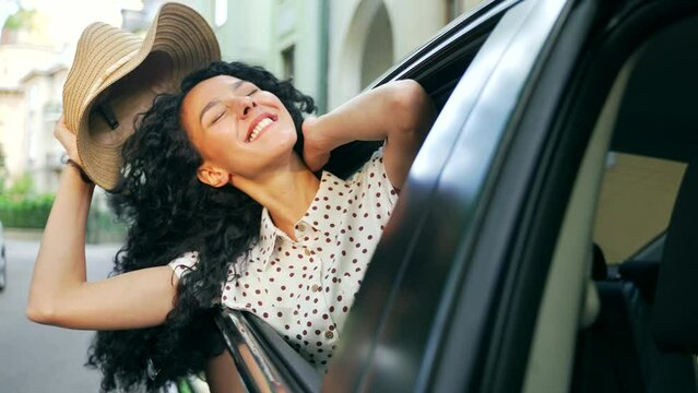 Portrait Of Young Pretty Hispanic Woman Sitting In Car And Leaning Out Of Passenger Side Car Window. Her Dark Fluttering Wind Rides Blows Hair. Concept Trip Journey Travelling Happy Girl On The Road