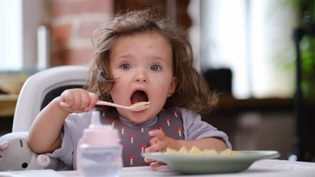 portrait caucasian baby girl about 2 years old in bib eating pasta from plate sitting high chair, self feeding for kids, baby-led weaning idea, healthy nutrition of solid food for infant