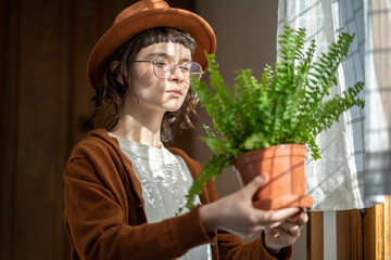 Plant lover. Hipster girl standing by window at home holding potted Boston fern houseplant, looking...