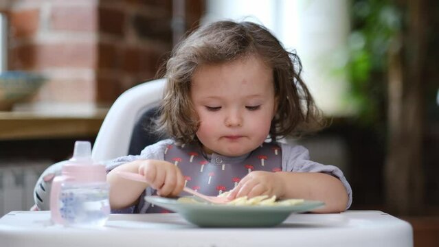 portrait caucasian baby girl about 2 years old in bib eating pasta from plate sitting high chair, self feeding for kids, baby-led weaning idea, healthy nutrition of solid food for infant