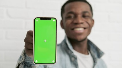 Happy young black man standing and pointing Big smartphone with blank green screen, demonstrating copy space for app or website design, standing over white brick background, mockup image