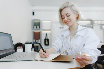 Young beautiful joyful blonde influencer woman blogger writes content plan in notepad in cafe with coffee, white light