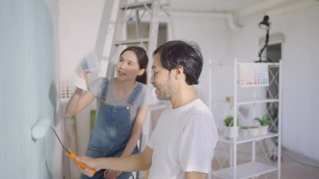 Young Asian couple repairing and painting the wall with blue paint using a roller during renovation in their new apartment.