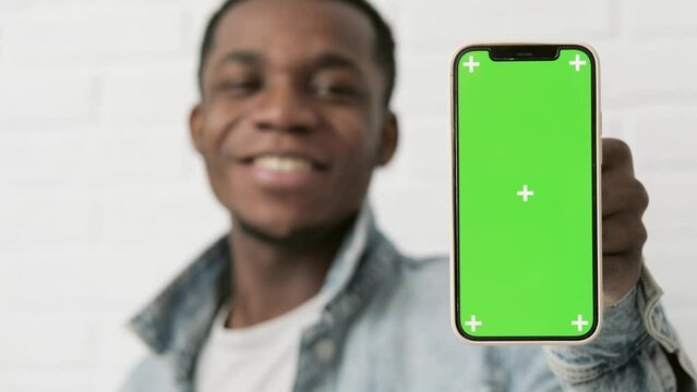 Happy young black man standing and pointing Big smartphone with blank green screen, demonstrating copy space for app or website design, standing over white brick background, mockup image