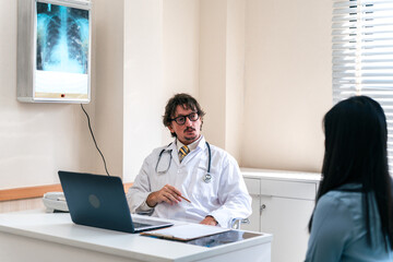 Fototapeta premium Caucasian doctor provides consultation and converses with an Asian female patient, while diagnosing the results of a chest X-ray and conducting an annual health check-up in workroom.