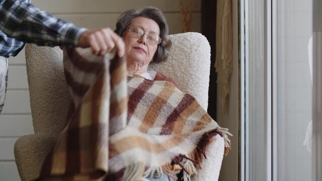 Elderly senior woman reading favorite bestseller novel poetry in paper book, enjoying cozy lazy weekend holiday time, relaxing on sofa at home. A loving husband snatches with a blanket.