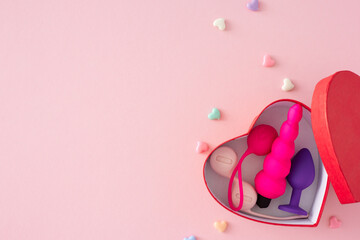 Erotic toys for adults gifts idea. Top view flat lay of red heart-shaped box, vaginal balls, anal plug, hearts decor on pastel pink background with space for greeting or ad