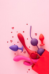 Concept of erotic pleasure toys. Top view vertical arrangement of shopping bag, vibrators, anal plugs, vaginal balls, red hearts on pastel pink background with empty space for text or advert