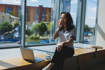 Fototapeta premium A young smiling curly-haired woman sits near the large windows in a cafe and works on a laptop. Remote work.