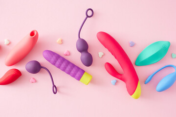 Satisfy your desires with a range of seductive pleasure toys. Top view flat lay of vibrators, massager for the clitoris, vaginal balls and colorful hearts on pastel pink background