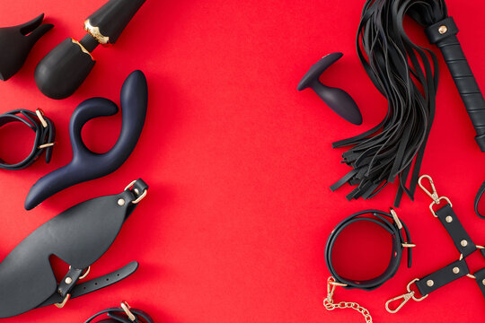 Erotic enjoyment BDSM toys idea. Top view flat lay of handcuffs, mask, leather whip, vibrator, dildo and anal plug on red background with empty space for promo or text