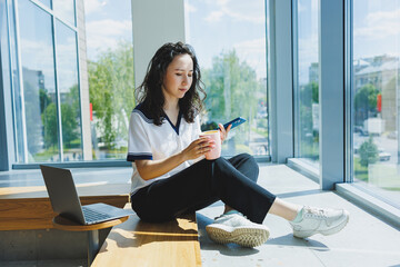Smiling curly-haired woman sitting in a loft cafe, working at a computer, reading messages on a smartphone. Remote work