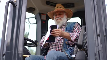 Portrait of happy senior farmer advertising Black Friday sales sitting in huge tractor. Front view confident male Caucasian agrarian looking at camera smiling showing advertisement in slow motion