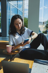 Freelance work. A young smiling curly-haired woman sits near the large windows in a cafe and works on a laptop. Remote work. A woman is talking on the phone while sitting in a cafe.