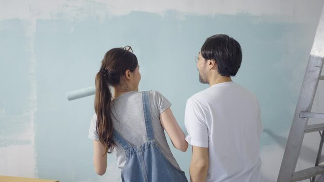 Young Asian couple Dancing together while repairing and painting the wall with blue paint using a roller during renovation in their new apartment.