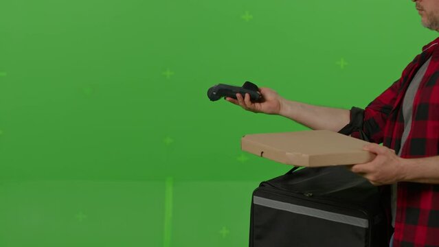 Caucasian male courier passing a box of pizza on green screen chroma key background. Man hold wireless bank payment POS terminal to process acquire credit card payment. Copy space mock up advertising