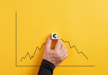 Predicting the market fluctuation in euro currency. Hand holds the wooden cube with euro symbol with an increasing and declining graph. - obrazy, fototapety, plakaty