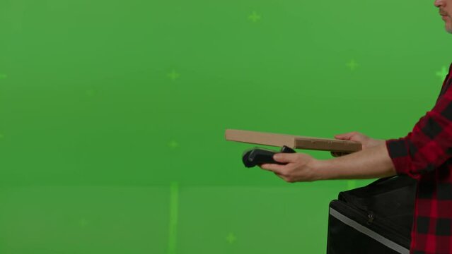 Caucasian male courier passing a box of pizza on green screen chroma key background. Man hold wireless bank payment POS terminal to process acquire credit card payment. Copy space mock up advertising