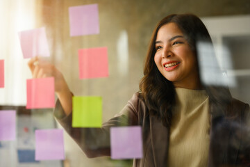 Cheerful asian female manager looking at paper memos on glass board, planning goals, manage corporate work tasks