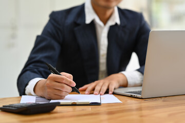 Cropped shot of male manager making important notes on paper, using laptop on wooden office desk