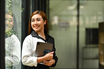 Portrait of cheerful young female entrepreneur holding digital tablet standing in office and looking away thoughtfully