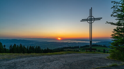farbiger Sonnenuntergang über dem Bodensee, mit einem Kreuz und Sitzbank im Vordergrund