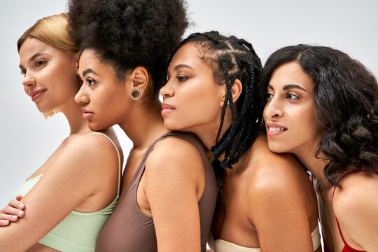Smiling and multiethnic women in colorful bras looking away while standing next to each other isolated on grey, different body types and self-acceptance concept, multicultural models