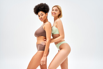 Smiling blonde woman in lingerie hugging african american friend and looking at camera isolated on grey, different body types and self-acceptance concept, multicultural models