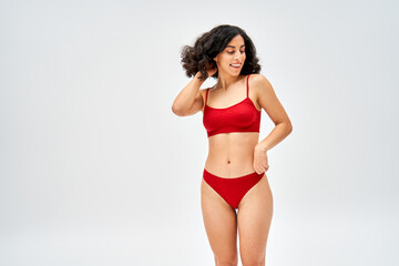 Smiling and brunette middle eastern model in red bra and panties touching hair and looking away while standing and posing isolated on grey, body acceptance concept