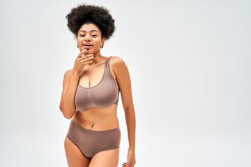Positive and seductive african american woman in brown panties and bra touching chin and looking at...