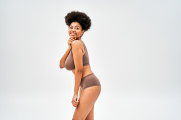 Cheerful african american model in brown lingerie touching chin and looking away while standing and...