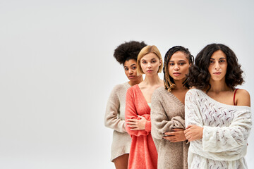 Multiethnic group of women in stylish knitted sweaters crossing arms and looking at camera isolated...