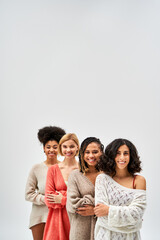 Positive multiethnic women in stylish knitted jumpers crossing arms and looking at camera while standing isolated on grey, different body types and self-acceptance, multicultural representation