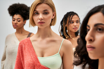 Obraz premium Portrait of young blonde woman in bra and wool jumper looking at camera while standing near blurred friends isolated on grey, different body types and self-acceptance, multicultural representation