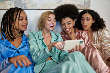 Excited blonde woman using blurred smartphone and sitting near cheerful multiethnic girlfriends in colorful pajama during girls night at home, bonding time in comfortable sleepwear, slumber party