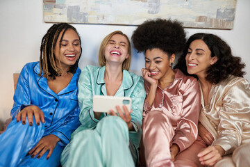 Obraz premium Young and laughing blonde woman holding blurred smartphone near multiethnic girlfriend in colorful pajama during girls night while sitting on bed at home, bonding time in comfortable sleepwear