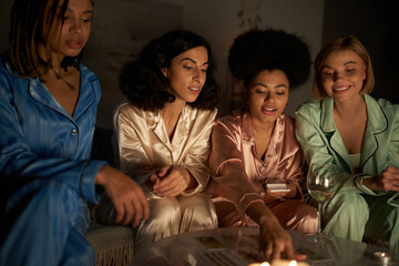 Fototapeta premium Smiling multiethnic girlfriends in colorful pajama looking at tarot cards on table near glass of wine and candles during girls night at home, bonding time in comfortable sleepwear, slumber party