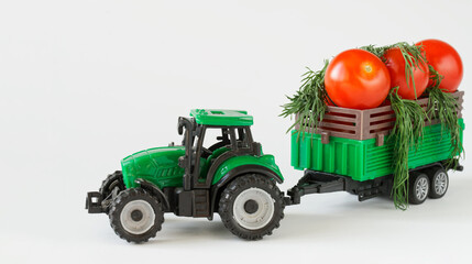 A toy tractor carries fresh red tomatoes and greens in a trailer. White background. The concept of agricultural work, harvesting and delivery of crops. Toy world.