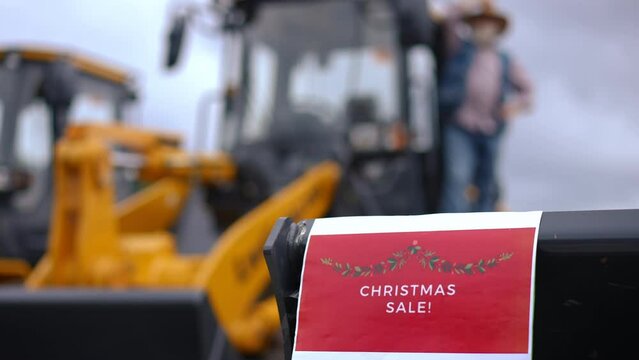 Christmas sale announcement on transport market with blurred man entering tractor cabin at background. Unrecognizable Caucasian senior farmer choosing new vehicle outdoors