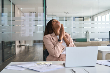 Euphoric professional young African American business woman executive feeling happy about financial work results, corporate goals achievement getting new job offer sitting with laptop in office.