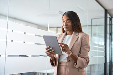 Young busy professional African American business woman company manager sales executive wearing suit holding tab using digital tablet computer standing in office managing financial data.