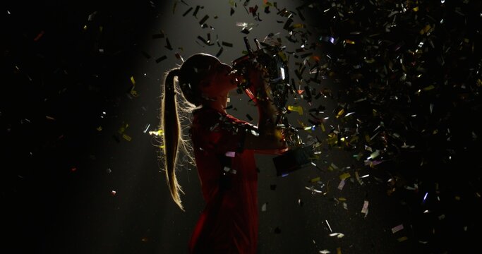 Silhouette of Caucasian woman female soccer football player raising a trophy above head against bright light and falling confetti. Super slow motion, shot on RED cinema camera