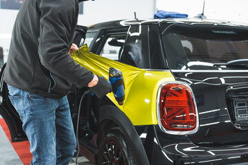 Unrecognisable car detailing specialist using heat gun to glue colourful car wrap to car paint. Indoor medium shot. High quality photo