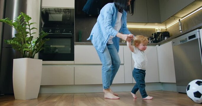 Mother helps the baby boy learn to walk. Child learns to take the first steps holding his mother's hands in modern kitchen at home. Happy family, mother support young toddler son.