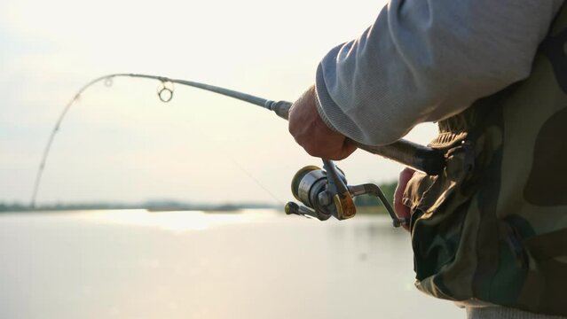 Old man fishing. Senior gray haired fisherman throws a spinning from shoreside at sunset, twisting a coil. Positive elderly male angling at lake, rotating reel rod. Retired fisher enjoying his weekend